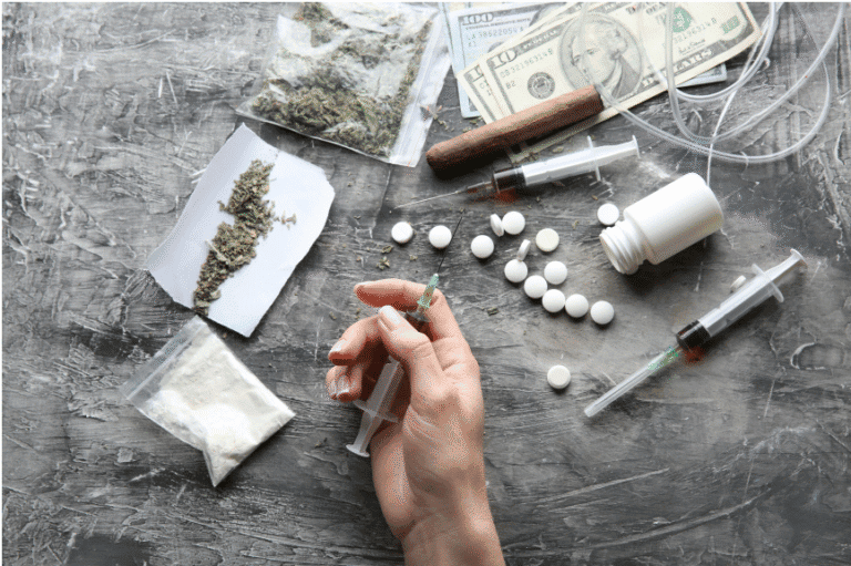 Hand preparing syringe surrounded by drugs, pills, and cash symbolising substance addiction.